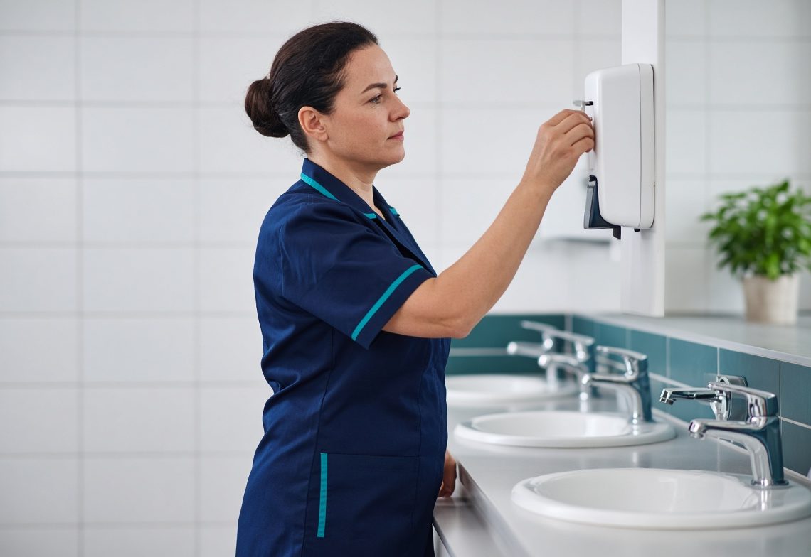 A cleaner refilling a soap dispenser in a gleaming washroom
